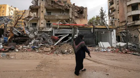 Reuters A woman dressed in a black headscarf and dark clothing walks past the wreckage of heavily damaged buildings in Beirut’s southern suburbs, Lebanon. The scene shows extensive destruction, with collapsed walls, twisted metal, and debris scattered across the ground. 