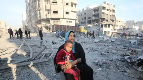 Getty Images A woman sits with a child as Palestinians inspect buildings damaged in an Israeli air strike in Gaza City, northern Gaza (14 January 2025)