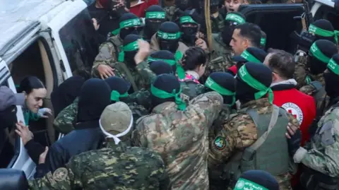 Reuters Doron Steinbrecher walks through a crowd of Hamas fighters, with their faces covered and wearing green bandanas, and Emily Damari gets out of a vehicle as both women are handed over to the Red Cross