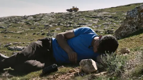 Dogwoof/Yabayay Media/Antipode Films Basel Adra lies on the grass in his home community of Masafer Yatta, his camera beside him, as an Israeli army bulldozer is seen on a rocky hill behind him 