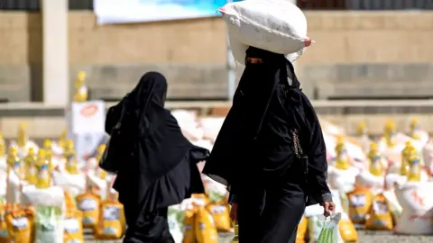 Getty Images A woman in black carries a sack of grain on her head. There are lots of other sacks in the background.