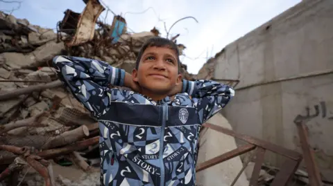 BBC Zakaria wears a blue and white hoodie as he sits on some rubble and puts his hands behind his head while smiling.