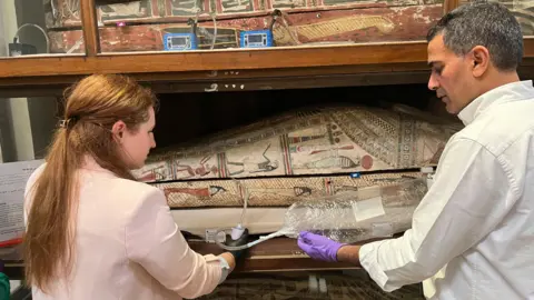 AP Two researches, dressed in white and wearing gloves, hold a tube into a sarcophagus. The tube is feeding into a bag of air. 