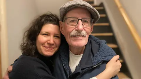 PA Media Sharone Lifschitz wears a black jumper and embraces her father, Oded Lifsthiz, who wears a hat and glasses