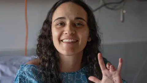 Emily Damari Family Emily Damari, a young woman with curly brown hair wearing a blue hospital gown, smiles and holds her hand to the camera while sat in a hospital bed, showing her two missing fingers.